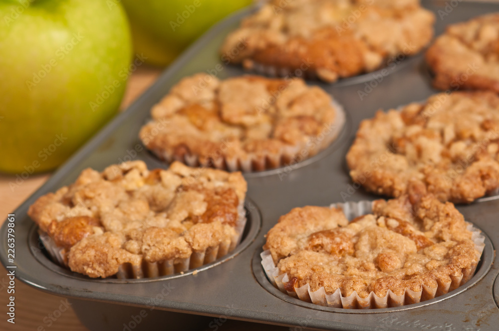 Top view, close up of six, freshly baked, homemade apple crumb muffins cooling in teflon, metal, cup cake baking sheet and two green apples Top view, close up of six, freshly baked, homemade apple crumb muffins cooling in teflon, metal, cup cake baking sheet and two green apples