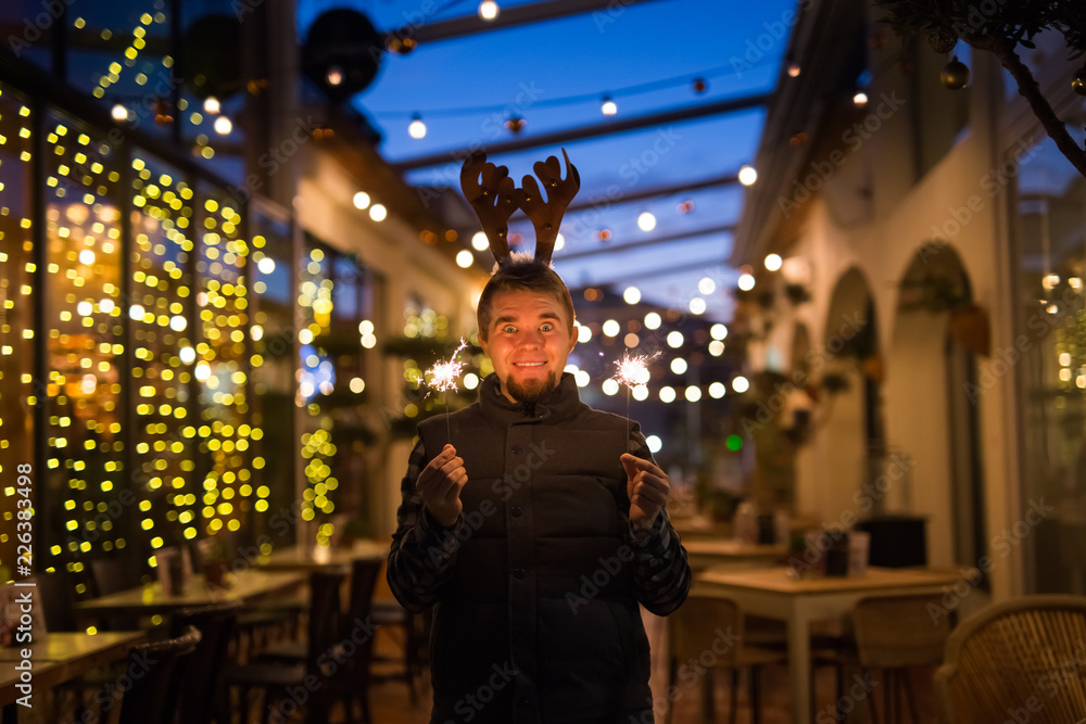 Fototapeta premium People, holidays and christmas concept - surprised man in deer's horns holding two bengals light or sparklers outdoors