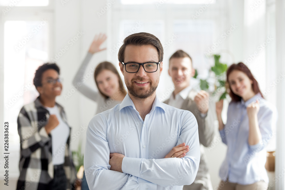 Smiling millennial confident businessman standing with arms crossed looking at camera with happy cheerful business partners team at background. Successful teamwork, business owner company boss concept Smiling millennial confident businessman standing with arms crossed looking at camera with happy cheerful business partners team at background. Successful teamwork, business owner company boss concept
