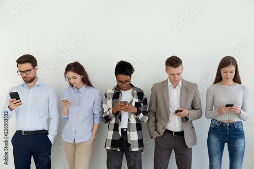Group of students or businesspeople standing in a row opposite white wall, holding looking on smartphones, waiting for interview. Chatting in social networks online addiction with devices concept