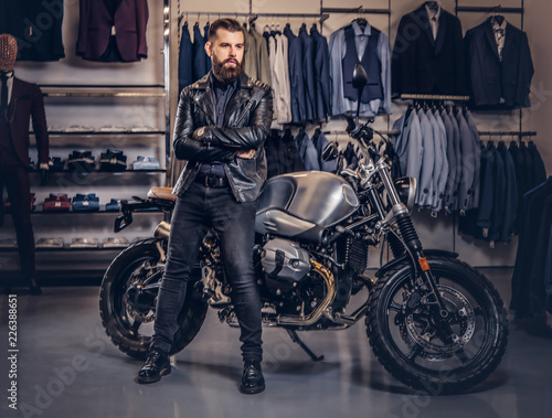 Brutal male dressed in black jacket leaning on a retro sports motorbike at the men's clothing store. © Fxquadro