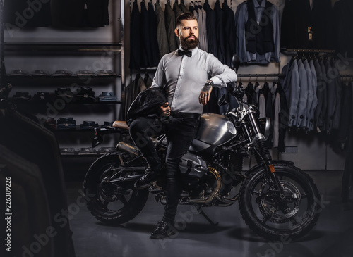 Elegantly dressed tattooed male with stylish beard holds black leather jacket posing near retro sports motorbike at the men's clothing store.