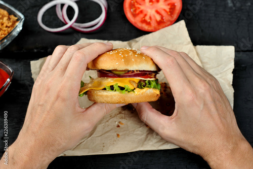 Cheeseburger in male hands. On the table are paper, onion rings, cups with ketchup and dried onions, a tomato. Black background. Close-up. View from above. 