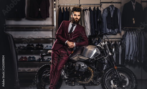 Handsome man with a stylish beard and hair dressed in vintage red suit posing near retro sports motorbike at men's clothing store.