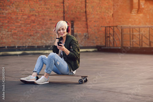 Fototapeta Naklejka Na Ścianę i Meble -  Pretty blond girl listening music in red headphones and using smartphone while sitting on the longboard at the background of brick wall.