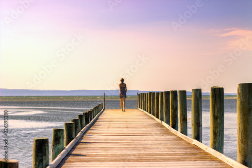Teen girl in a dress walking along the pier towards the sea. Early summer morning.