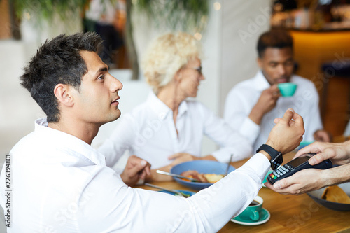 Serious handsome young manager in white shirt sitting at table with friends and paying for lunch with smartwatch in restaurant