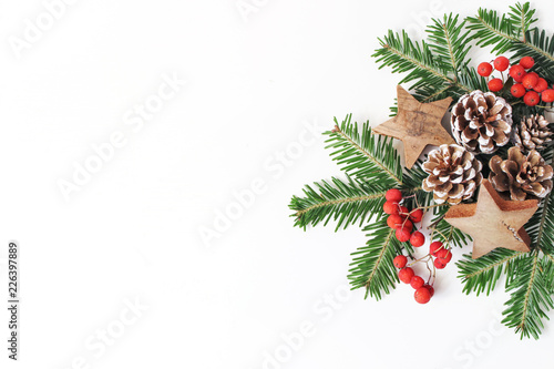 Christmas festive styled floral composition. Pine cones, fir tree branches, red rowan berries and wooden stars on white table background. Decorative frame, web banner. Flat lay, top view. Copy space.