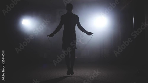 Boxer does some jump rope exercises in dark room under light. Fighter training in smoky studio in slow motion. Silhouette on dark background photo