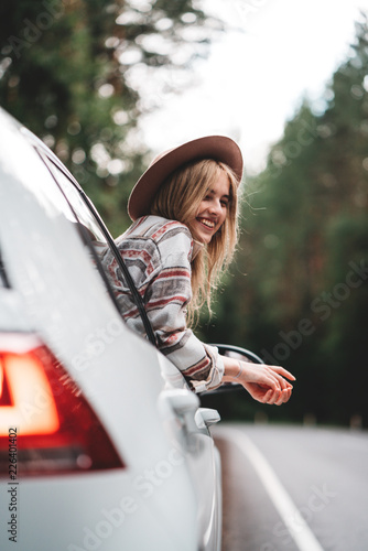 Hipster woman traveling by car on wild forest road. Handsome happy girl wearing checkered shirt and hat is sitting at the wheel of a car and looking from window. Wanderlust photo