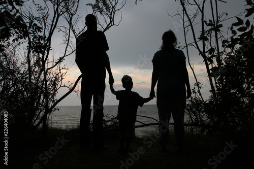 Silhouette of a man, a woman and a small child on the background of a sea sunset. Family with a baby are standing on the seashore in a frame of bushes, black silhouette