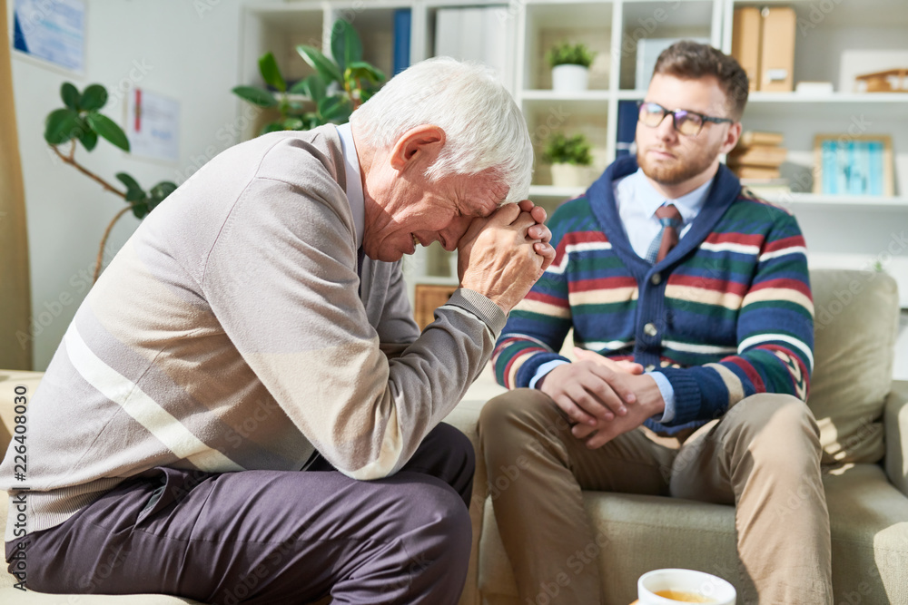 Sad emotional senior patient sitting on sofa and leaning on hands while crying during psychotherapy session, attentive psychiatrist listening to him