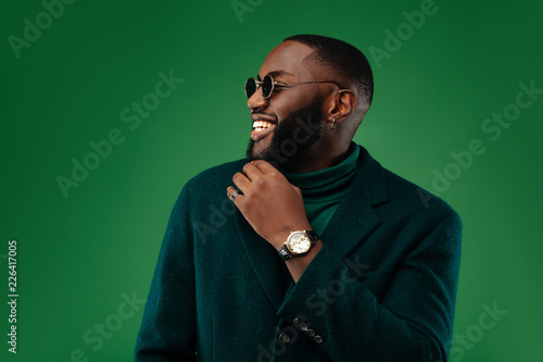 Man portrait. Style. Handsome Afro American guy in green jacket and sun glasses is smiling, on a green background