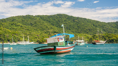 Colorful fishing wooden boat at a bay surrounded by other ships, warm clear blue sea water and a green tropical jungle behind, at a beautiful summer vacation island, Ilha Grande, Brazil