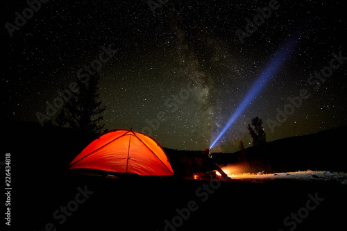 A tourist watches the starry sky near the tent on the background of a beautiful and winding river.