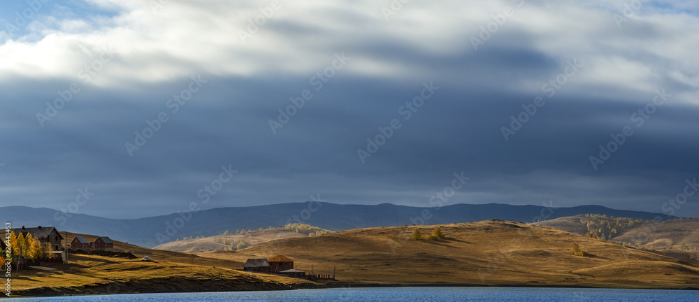 Obraz premium Wide panoramic photo of countryside landscape in autumn day with hill slopes illuminated by sunlight shining through the cloudy sky; meadows on sunset and long shadows of trees; yellow and blue shades