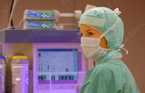 A young woman poses in an operation theater fully dressed as a anesthetic theater nurse with a face mask and green sterile medical work clothing.