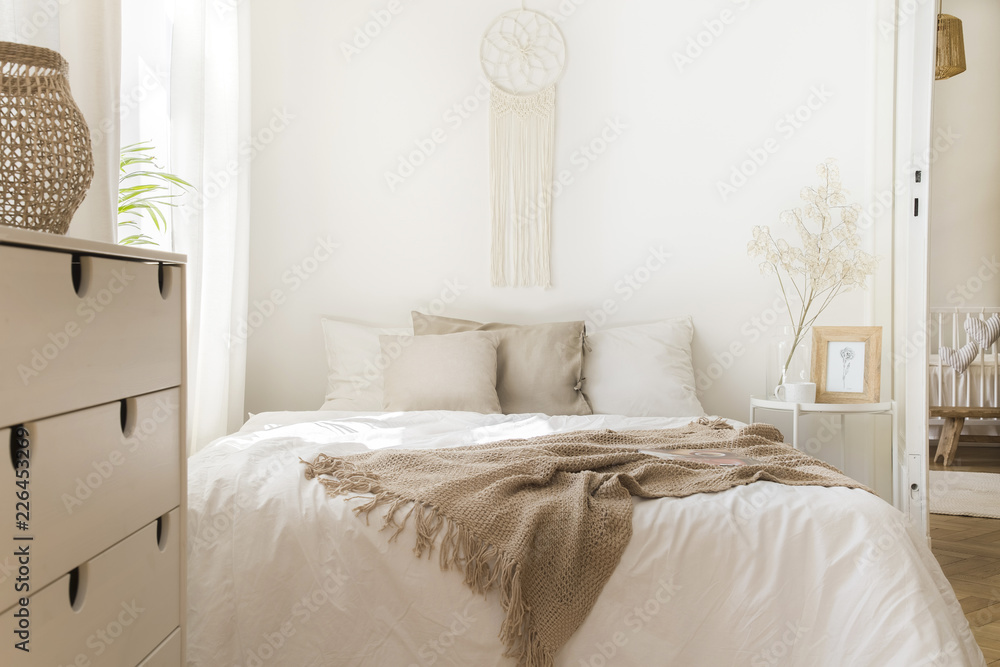 Fototapeta Koc na białym łóżku z poduszkami i drewnianą szafką w minimalistycznym naturalnym wnętrzu sypialni. Prawdziwe zdjęcie