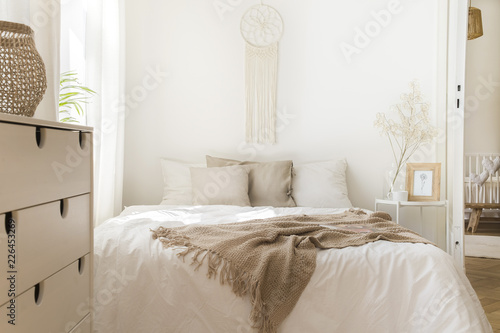 Blanket on white bed with pillows and wooden cabinet in minimal natural bedroom interior. Real photo