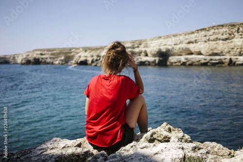 Young woman sitting on the top of rock and looking at the seashore and mountains at colorful sunset in summer. Landscape with girl, sea and mountain. Russia, Crimea.