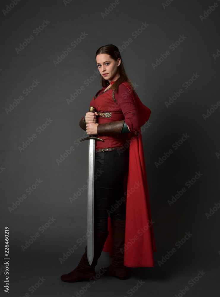  full length portrait of brunette girl wearing red medieval costume and cloak. standing pose  holding a sword on grey studio background.