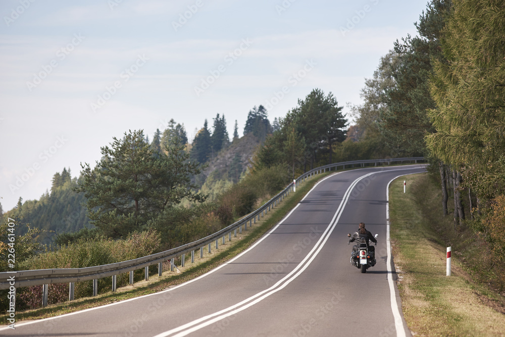 Back view of motorcyclist riding motorbike up twisted empty asphalt road on bright sunny summer day by green forest trees under clear blue cloudless sky.