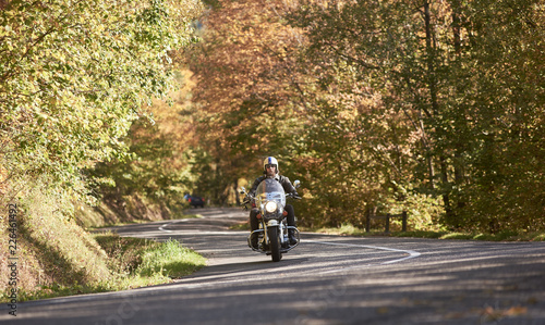 Wallpaper Mural Bearded motorcyclist in helmet, sunglasses and black leather clothing riding motorbike along empty asphalt road winding among tall green trees on bright sunny autumn day. Active lifestyle concept. Torontodigital.ca