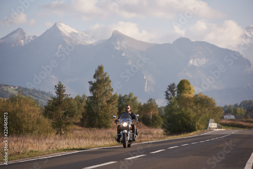 Bearded motorcyclist in sunglasses and black leather clothing riding cruiser powerful high-speed motorbike on blue foggy mountains with white snowy peaks and bright summer sky copy space background.