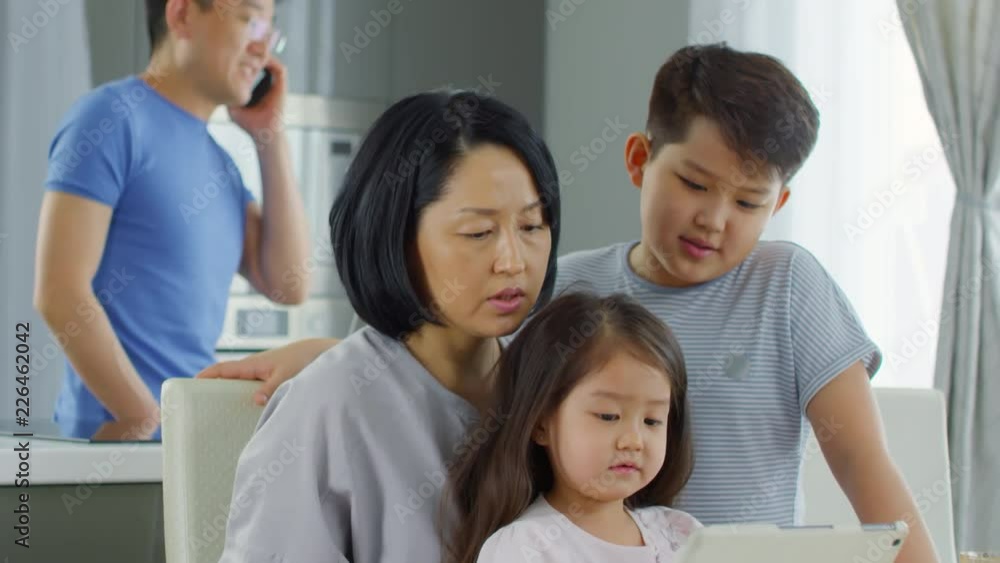 Tilt down shot of Asian mother, cute little girl and school-age boy chatting and playing on tablet in kitchen as middle-aged man in glasses talking on mobile phone in background Tilt down shot of Asian mother, cute little girl and school-age boy chatting and playing on tablet in kitchen as middle-aged man in glasses talking on mobile phone in background