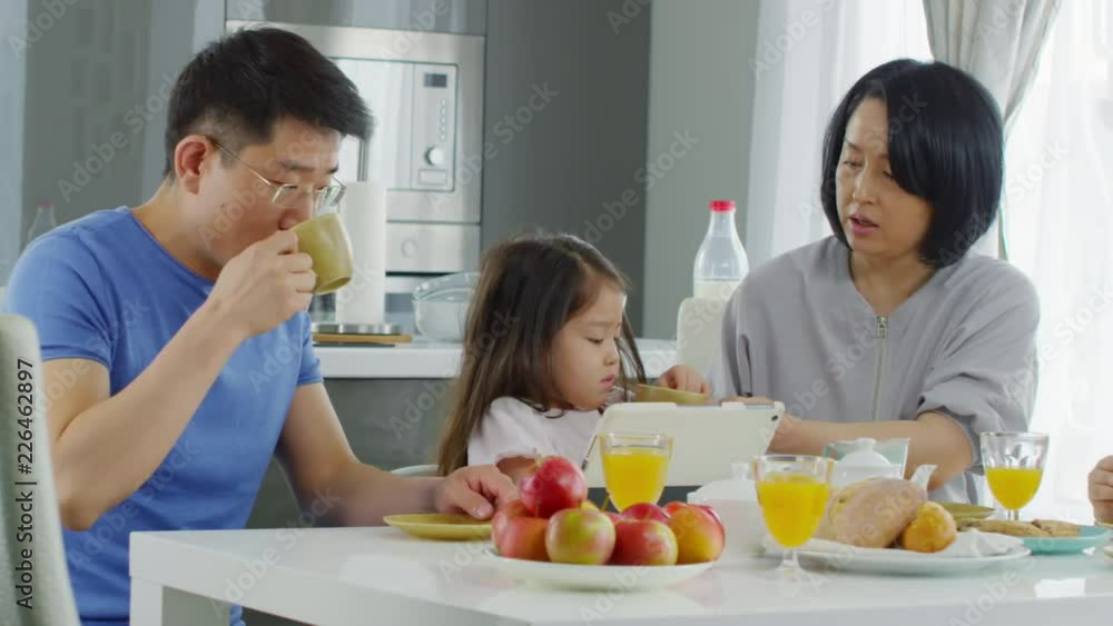 PAN of happy Asian family having breakfast in morning: father in glasses talking and drinking coffee as mother giving cup of milk to cute little girl. School-age boy sitting beside them and smiling PAN of happy Asian family having breakfast in morning: father in glasses talking and drinking coffee as mother giving cup of milk to cute little girl. School-age boy sitting beside them and smiling