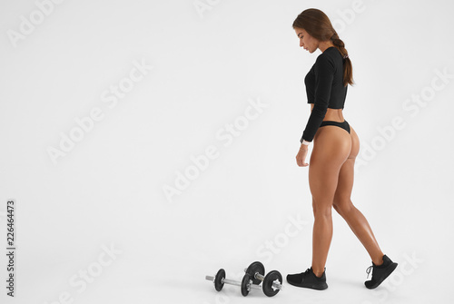 Indoor picture of gorgeous athletic woman with braid posing in studio wearing thongs, top and running shoes during morning training, doing exercise for butt muscles using dumbbells on floor