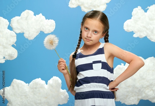 girl has a big dandelion in her hands, dressed in striped dress, posing on a blue background with cotton clouds, the concept of summer, holiday and happiness