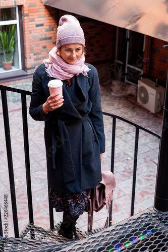 Young attractive lady in pink knitting hat and black coat drink coffee to go . Smiling beautiful hipster happy woman in city street, wearing stylish clothes. Autumn trend, urban stydent style. photo