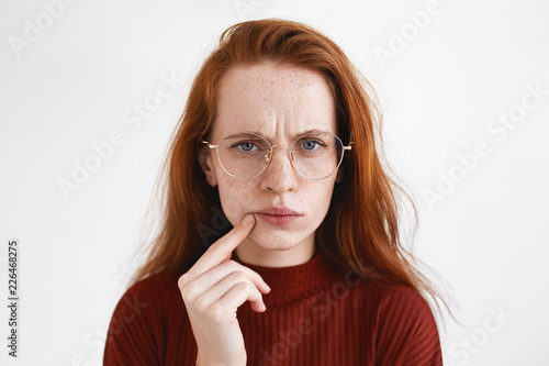 Picture of puzzled young red haired woman in spectacles frowning, having suspicious look, touching lips. Beauitful pensive ginger girl being frustrated with something. Human facial expressions