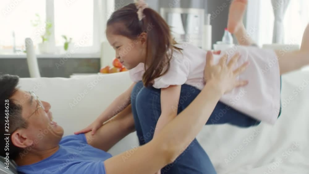 Handheld medium shot of Asian middle-aged father in glasses lying on sofa and lifting cute preschool-age girl with his legs Handheld medium shot of Asian middle-aged father in glasses lying on sofa and lifting cute preschool-age girl with his legs