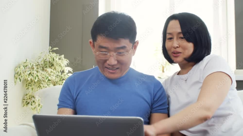 Handheld tracking shot of Asian woman and man sitting on sofa and discussing something while looking at laptop Handheld tracking shot of Asian woman and man sitting on sofa and discussing something while looking at laptop
