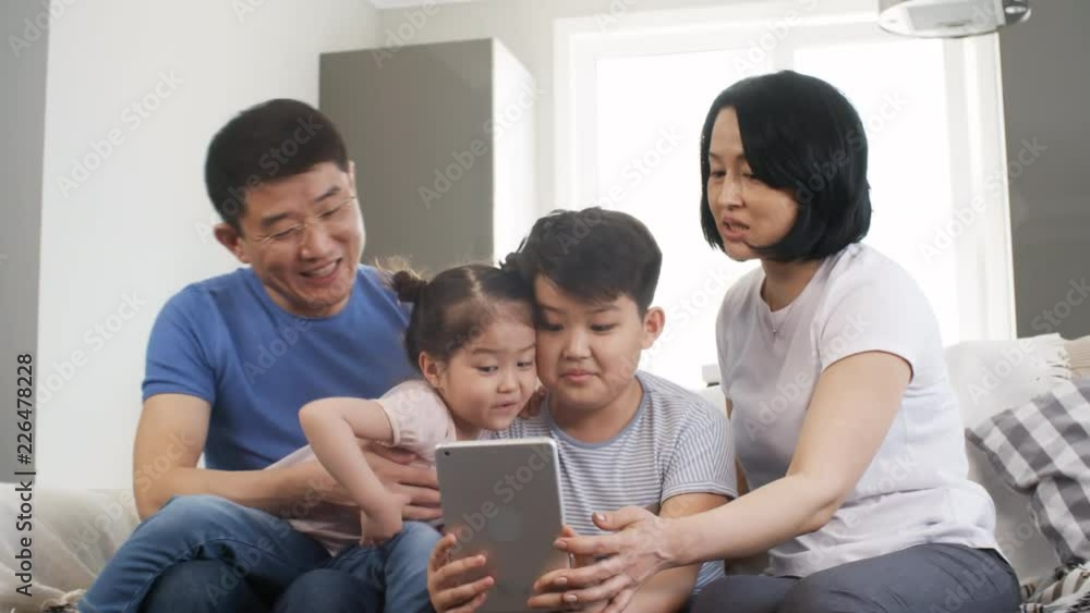 Tracking shot of happy Asian parents and children sitting on sofa and playing with tablet: they are laughing and taking selfies together Tracking shot of happy Asian parents and children sitting on sofa and playing with tablet: they are laughing and taking selfies together