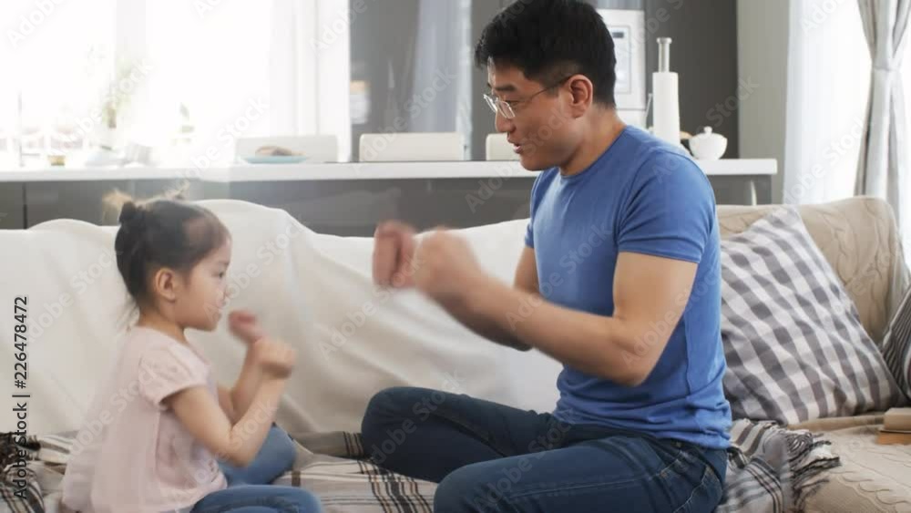 Tracking shot of cheerful Asian middle-aged father in glasses and cute little girl sitting on sofa and playing rock-paper-scissors Tracking shot of cheerful Asian middle-aged father in glasses and cute little girl sitting on sofa and playing rock-paper-scissors