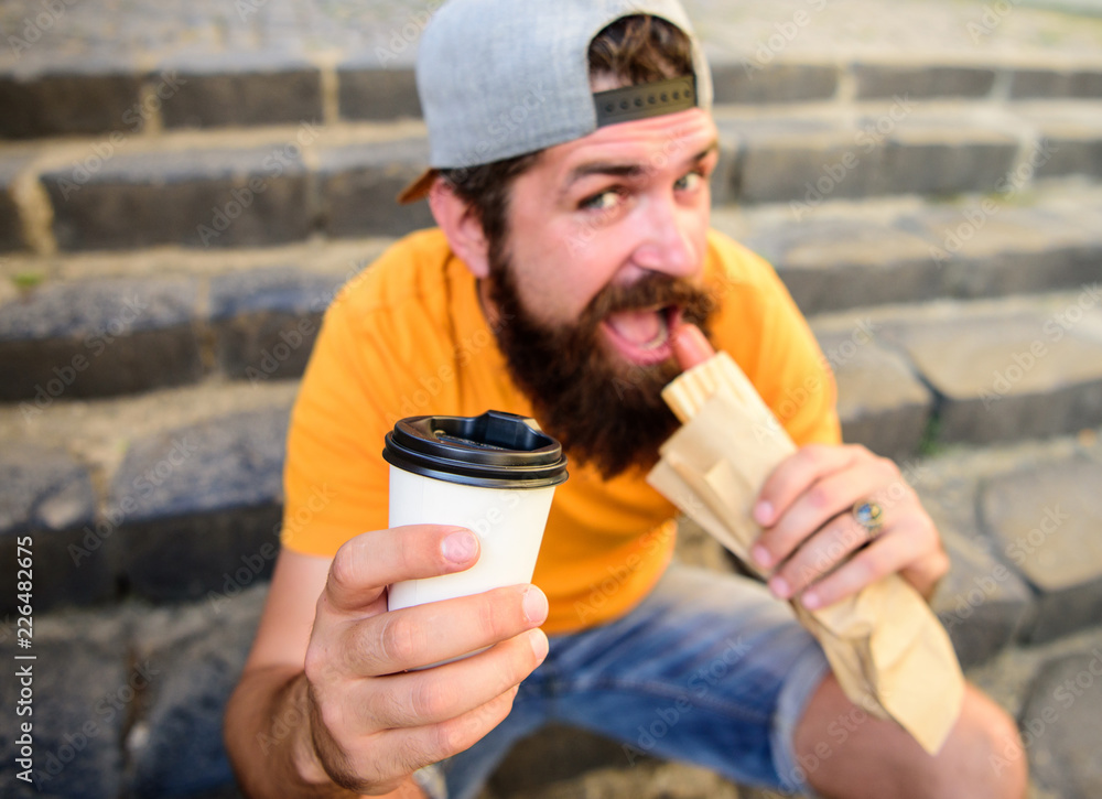 Man bearded enjoy quick lunch stairs background. Energy from street food daily traditional snack. Hipster bite eat hot dog. Fast food meal for lunch. Hipster bite hot dog hold drink paper cup Man bearded enjoy quick lunch stairs background. Energy from street food daily traditional snack. Hipster bite eat hot dog. Fast food meal for lunch. Hipster bite hot dog hold drink paper cup