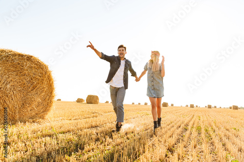 Photo of young couple man and woman walking through golden field, with haystacks during sunny day