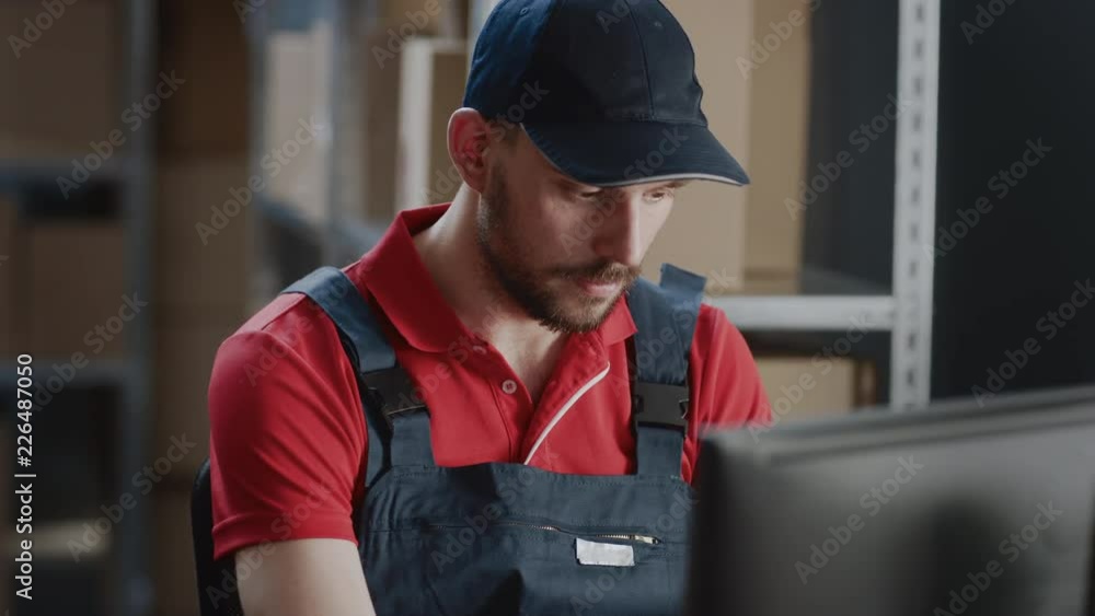 Portrait of Uniformed Worker Using Personal Computer while Sitting at His Desk in the Warehouse. In the Background, Room with Shelves Full of Cardboard Box Packages Ready For Shipping. Portrait of Uniformed Worker Using Personal Computer while Sitting at His Desk in the Warehouse. In the Background, Room with Shelves Full of Cardboard Box Packages Ready For Shipping.