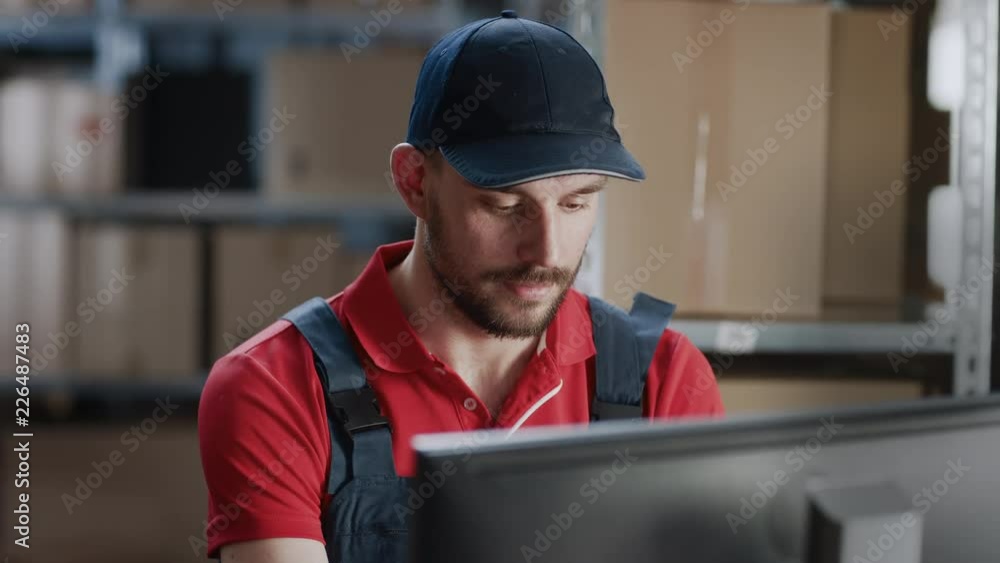 Portrait of Uniformed Worker Using Personal Computer while Sitting at His Desk in the Warehouse. In the Background, Room with Shelves Full of Cardboard Box Packages Ready For Shipping. Portrait of Uniformed Worker Using Personal Computer while Sitting at His Desk in the Warehouse. In the Background, Room with Shelves Full of Cardboard Box Packages Ready For Shipping.