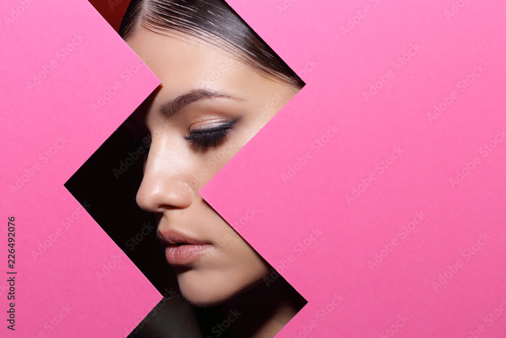 Fototapeta premium Twarz młoda piękna dziewczyna z jaskrawym makijażem patrzeje przez dziury w różowym papierze