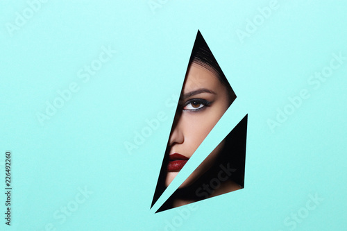 Face of young beautiful girl with a bright make-up and red lips looks through a triangle hole in green paper.