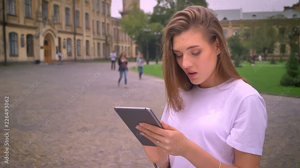awesome blond caucasian woman is typing in her tablet with concentrated face while standing on the street with buildings on background awesome blond caucasian woman is typing in her tablet with concentrated face while standing on the street with buildings on background