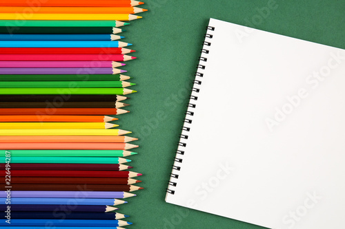 Top view of a set of many colorful pencils and empty sketchbook with a copy space on a green background. Art, craft, school and education concept. photo