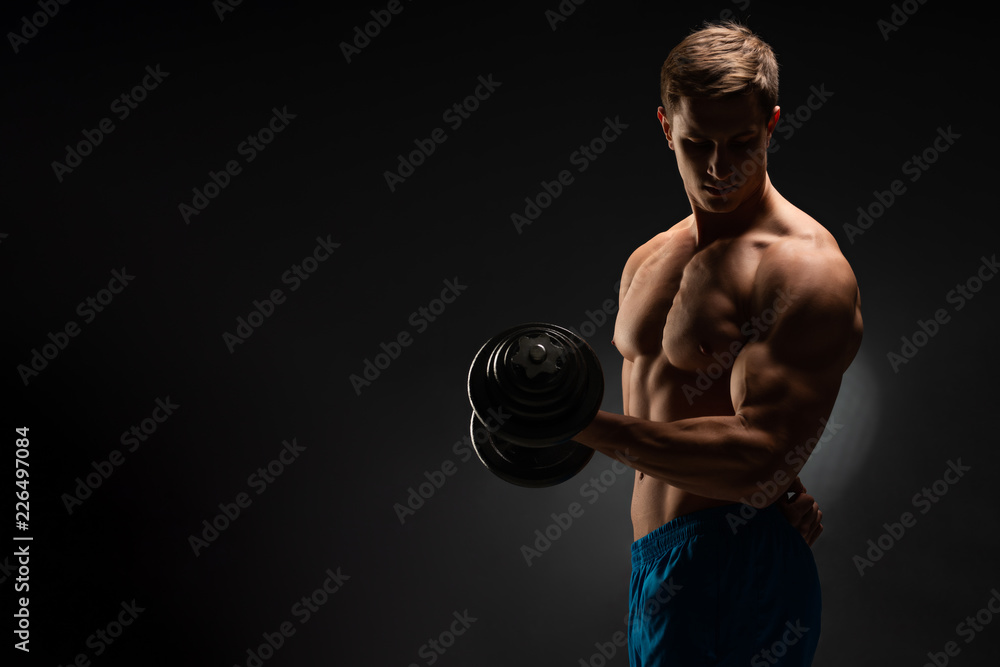 Handsome power athletic man in training swap muscles with dumbbells. A strong six-pack bodybuilder, perfect abs, shoulders, biceps, triceps and chest. On a dark background under the banner