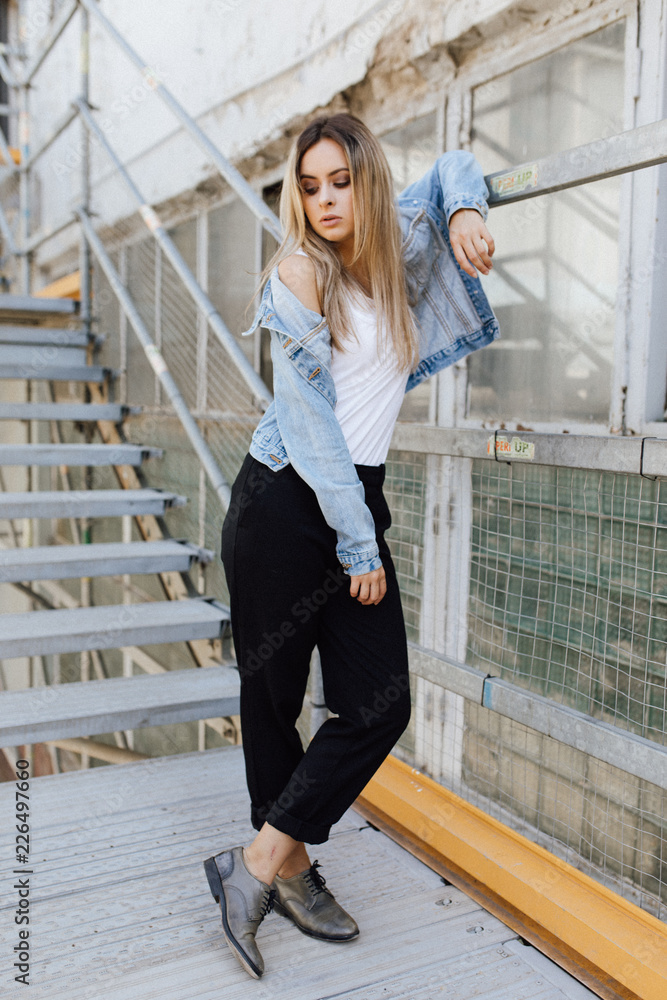 Charming young blonde woman dressed in casual style poses before concrete footsteps in an industrial building