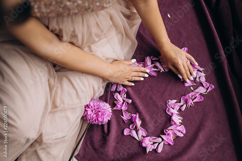 Pregnant woman in golden dress makes a heart of pink petals sitting on violet plaid