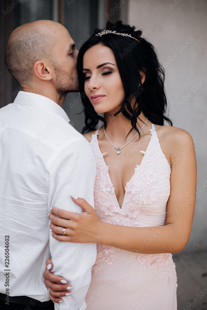 Handsome young bald headed man hugs with passion young brunette in pink evening dress Handsome young bald headed man hugs with passion young brunette in pink evening dress